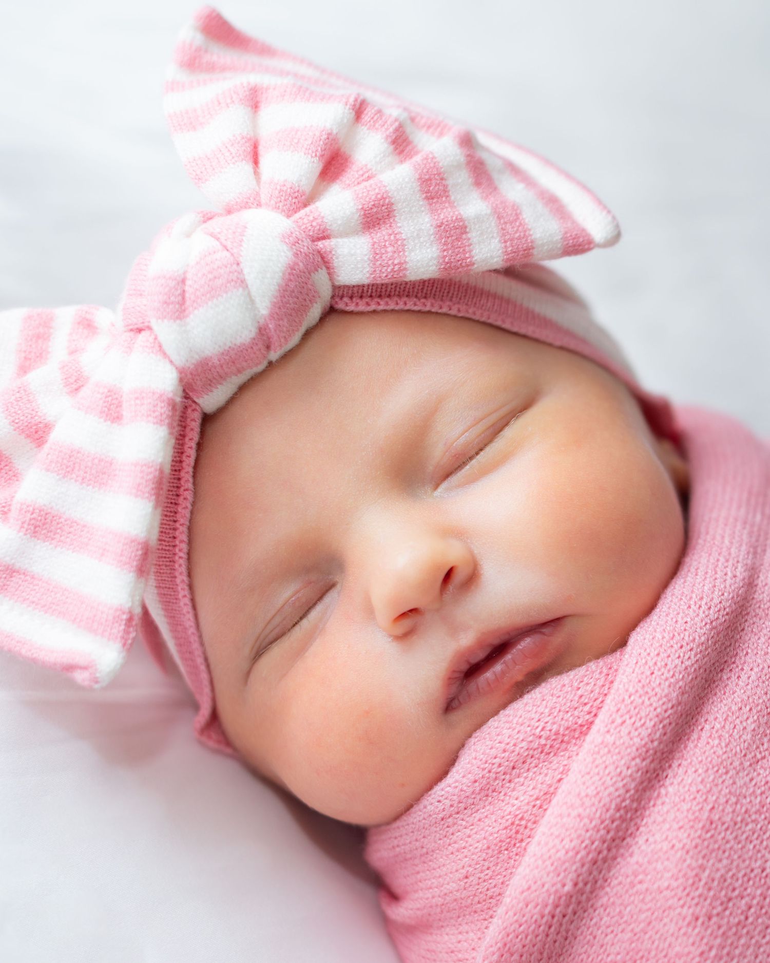 A sleeping newborn baby wearing a pink knit outfit and striped hat rests peacefully against a white background.