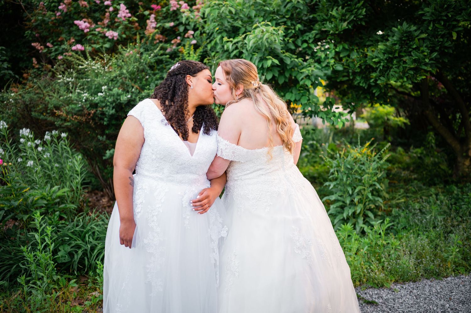 Two brides in white gowns share an intimate moment in a garden setting with flowers.