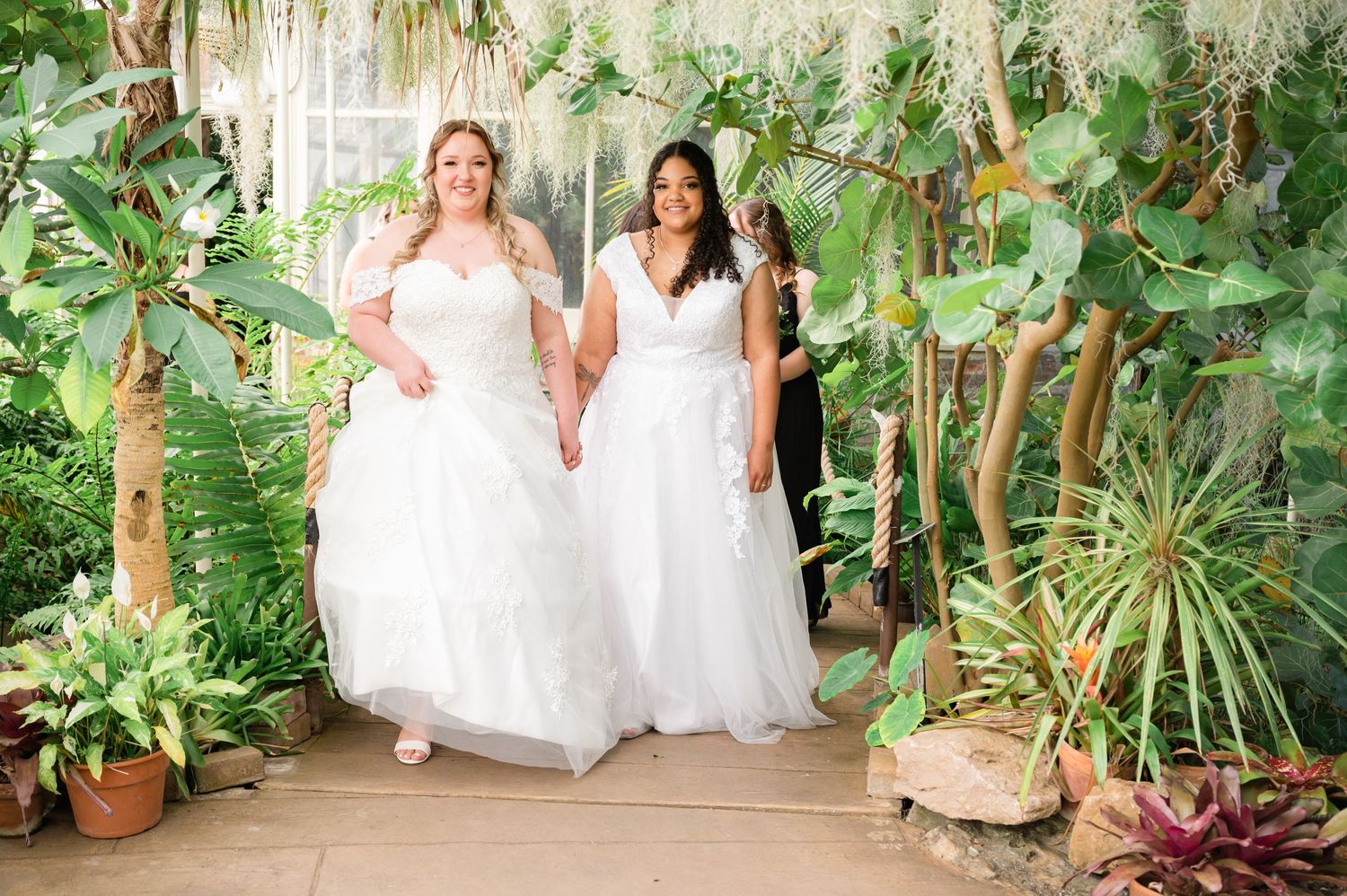 Two brides in white dresses walk through a lush greenhouse filled with tropical plants.