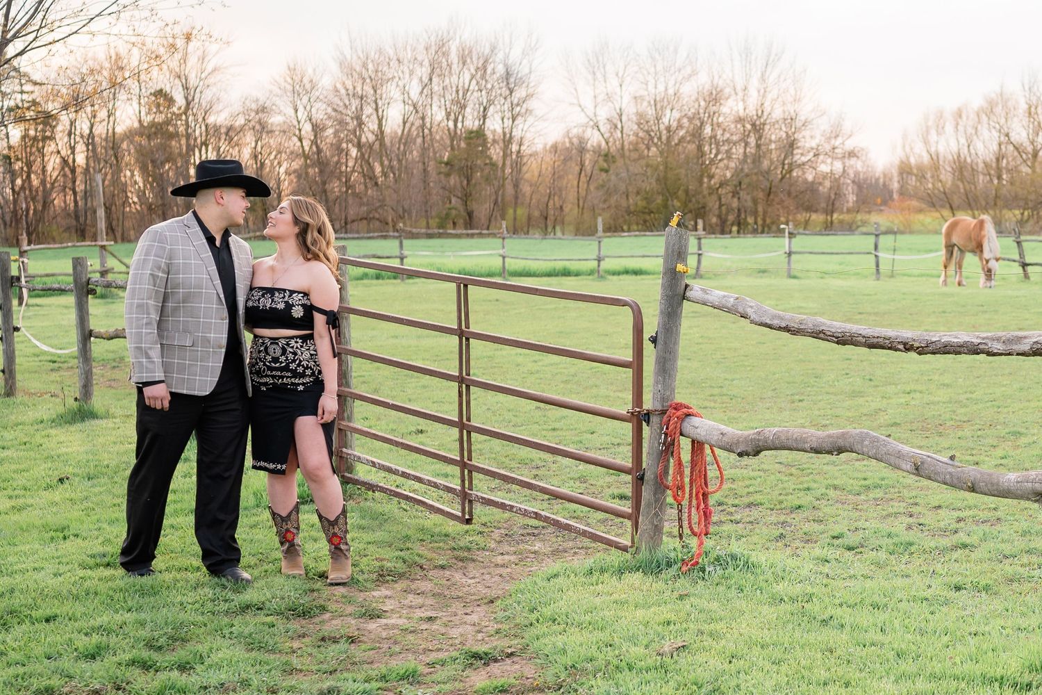 Stylishly dressed pair stand together by rustic gate with horses visible in background.