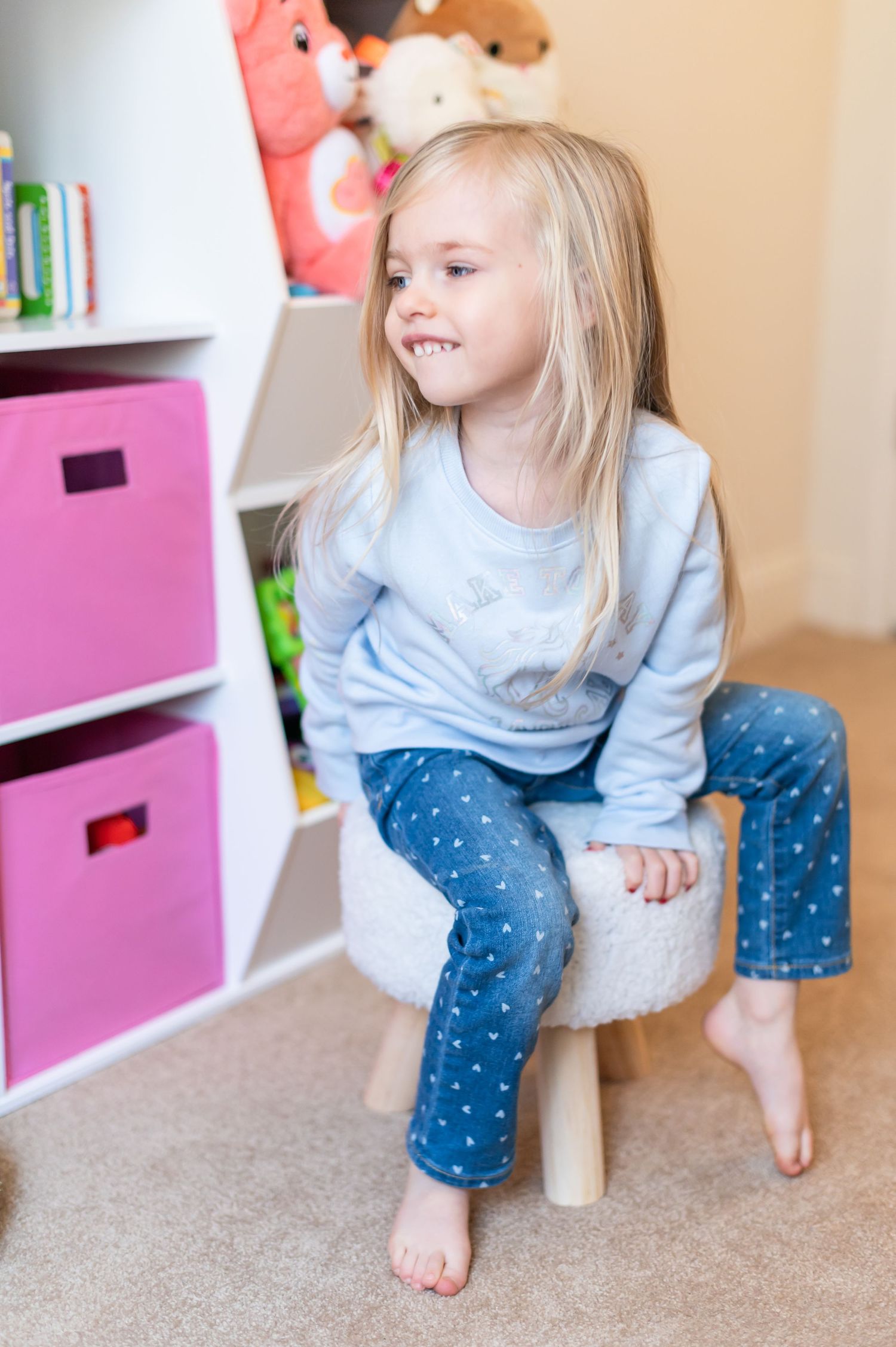 Child sitting on a white stool in a playroom with pink storage boxes and stuffed animals.