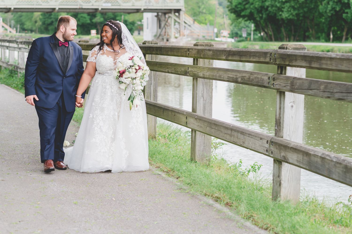Wedding couple walks along waterside path with wooden railing, surrounded by greenery.