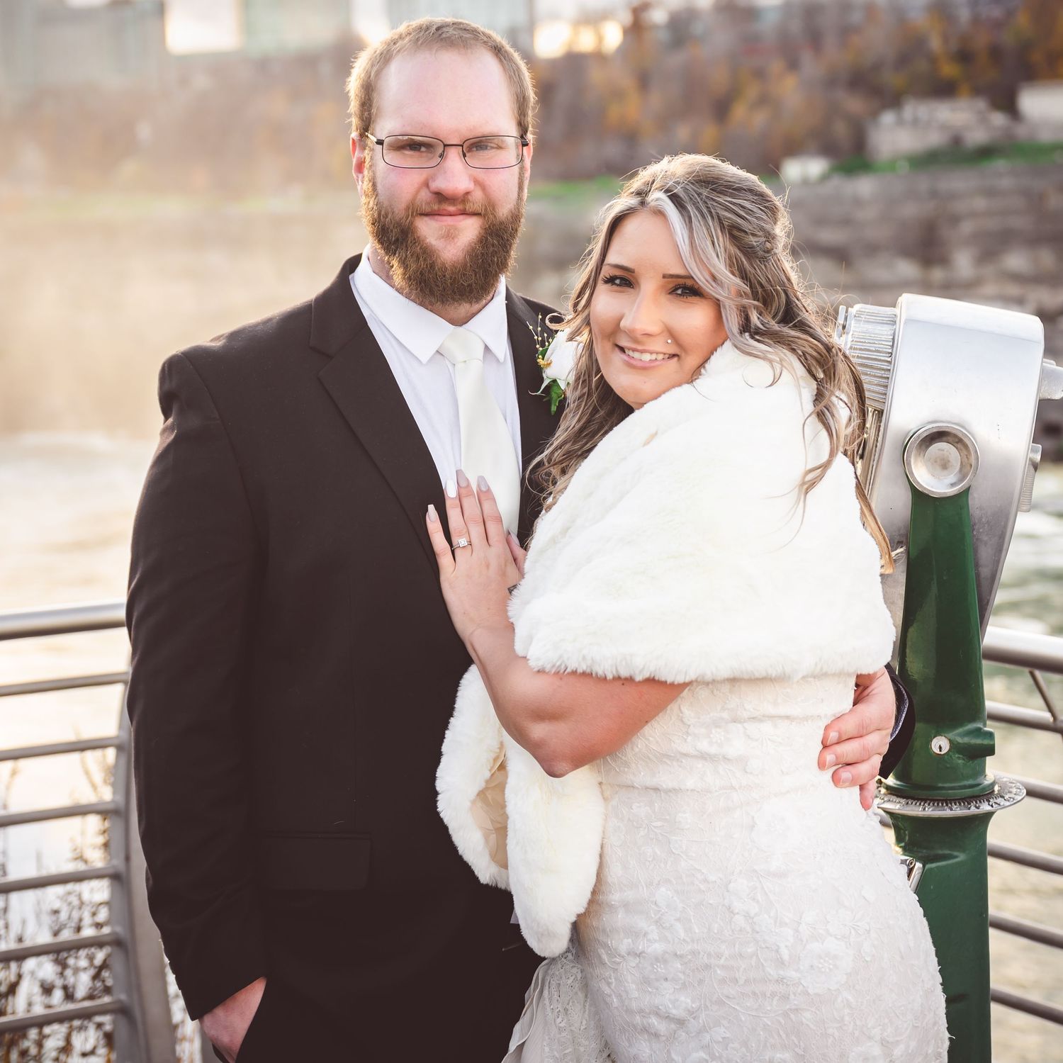 Newlyweds embrace on riverside overlook during autumn sunset with cityscape in background.