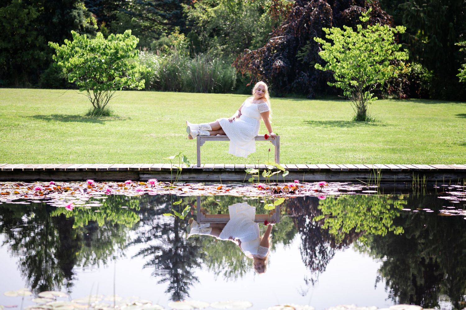 A serene pond reflects trees and sky while surrounded by lush summer greenery.