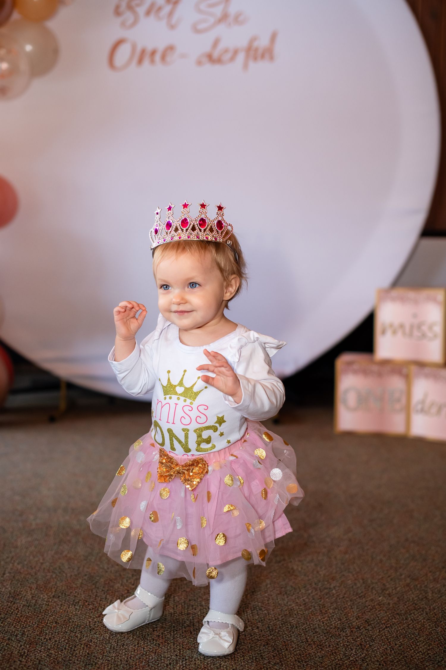 A small child wearing a pink tulle skirt and white top celebrates their first birthday with a pink crown and party decor.