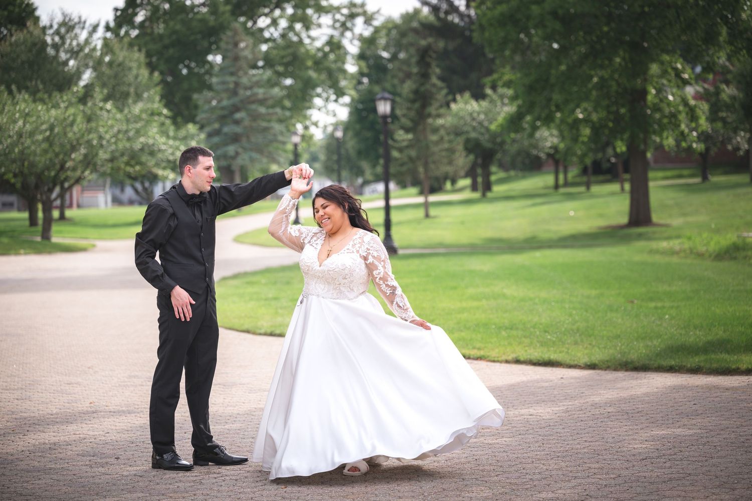 A couple in formal wedding attire dance playfully on a path in a lush green park during their special day.