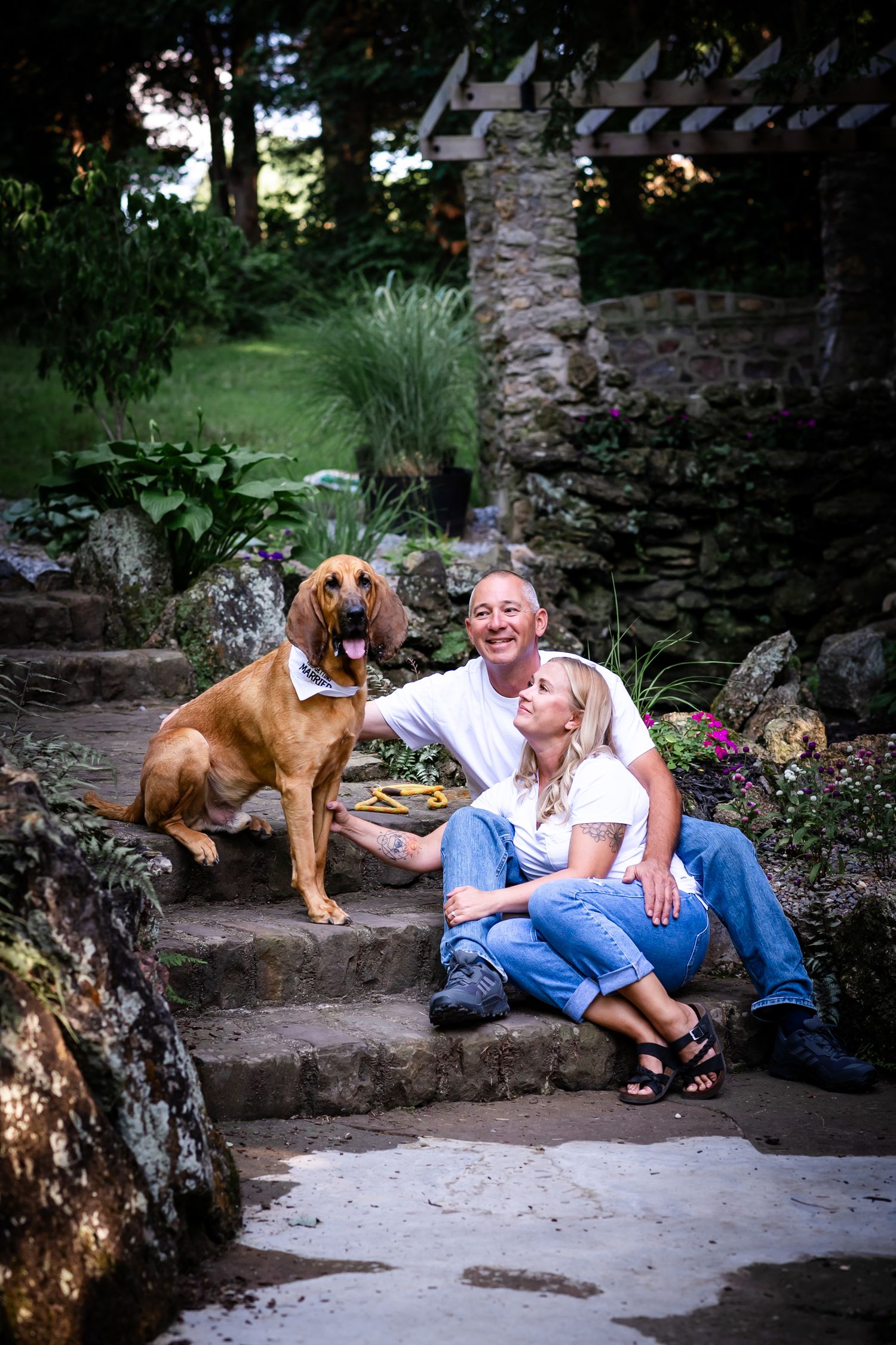 A couple relaxes on stone steps with their brown bloodhound dog in a garden setting at dusk.