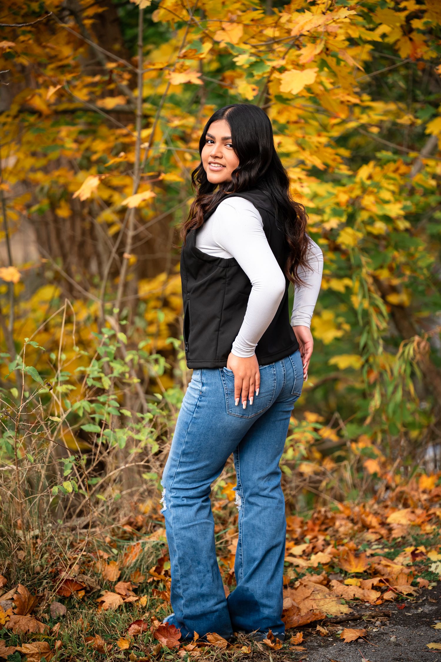 Person in blue jeans and white top poses against vibrant autumn foliage with golden yellow leaves in background.