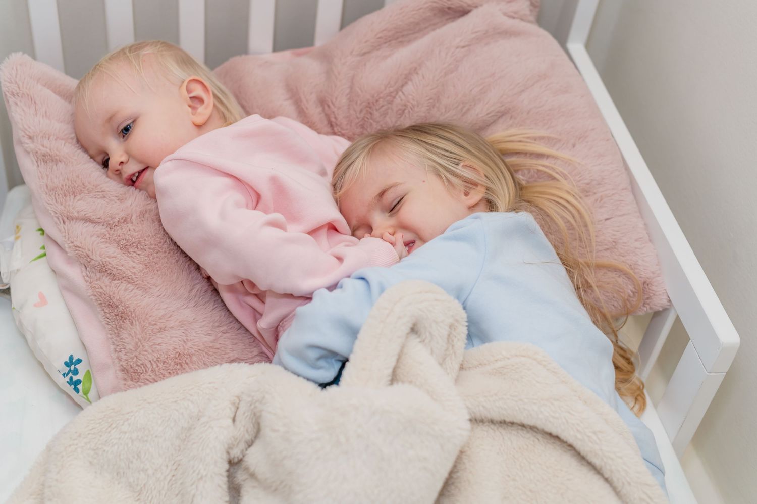 Two young children sleeping peacefully together under cozy blankets in a white crib.