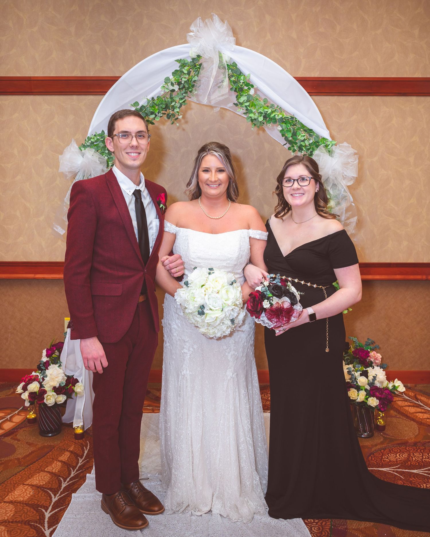 Three people pose under a wedding arch, with the bride in white between two attendants in burgundy and black.