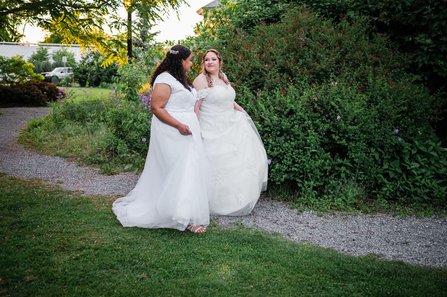 Two wedding gowns on a garden path with lush greenery in the background.