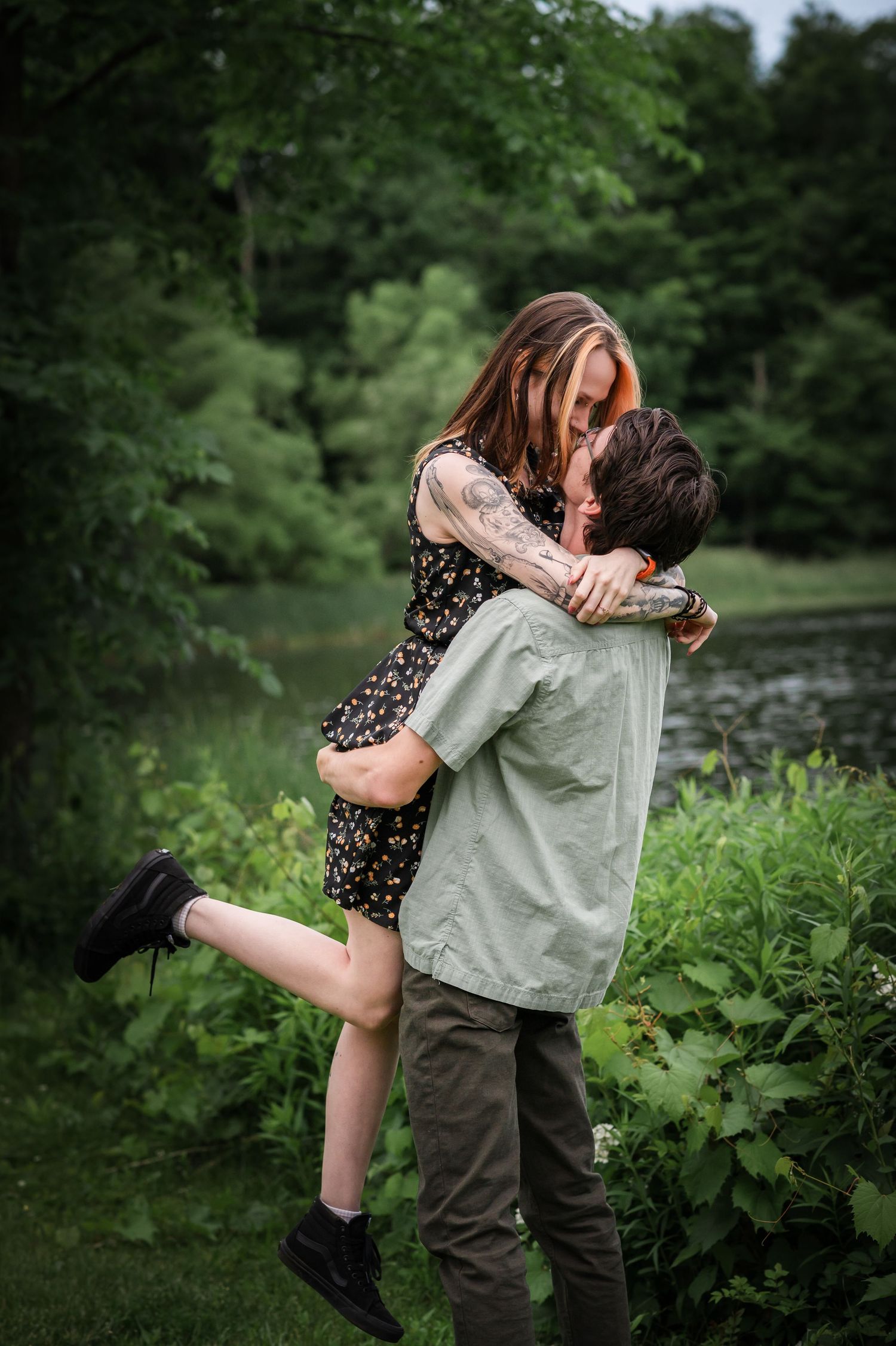 A romantic couple embraces by a serene lake surrounded by lush greenery on a summer day.