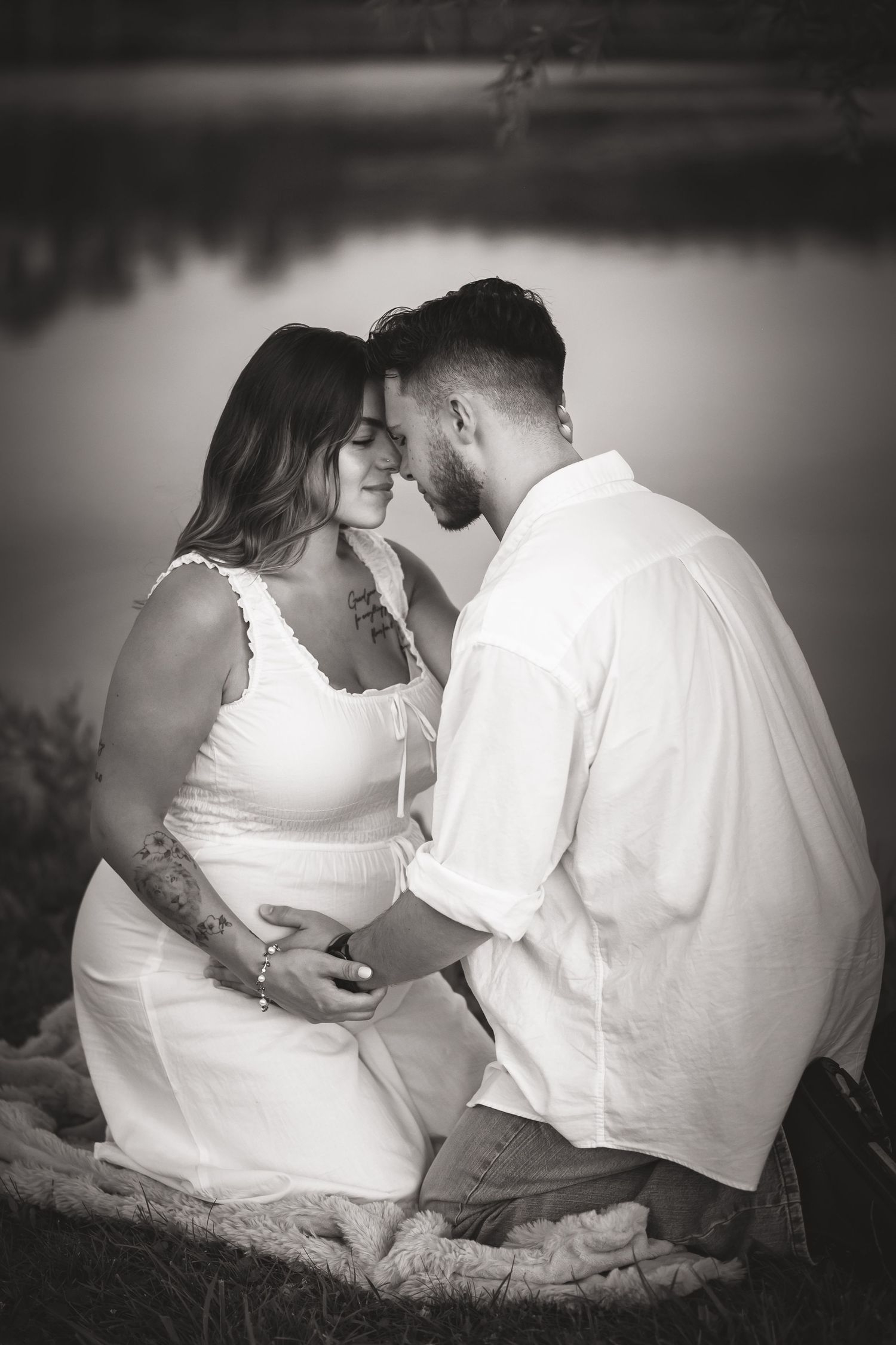 Black and white photo of a couple in white outfits sharing an intimate moment by a lake, cradling a baby bump.