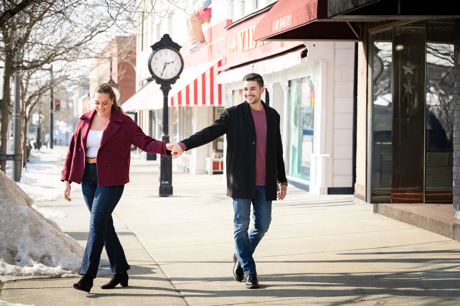 Couple holding hands walking on a sunny small-town sidewalk in winter coats near a street clock.