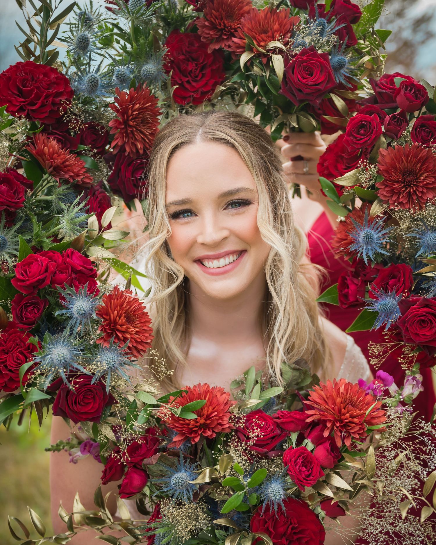 Vibrant burgundy and deep red flowers with blue thistle create a dramatic floral arrangement with rustic greenery accents.