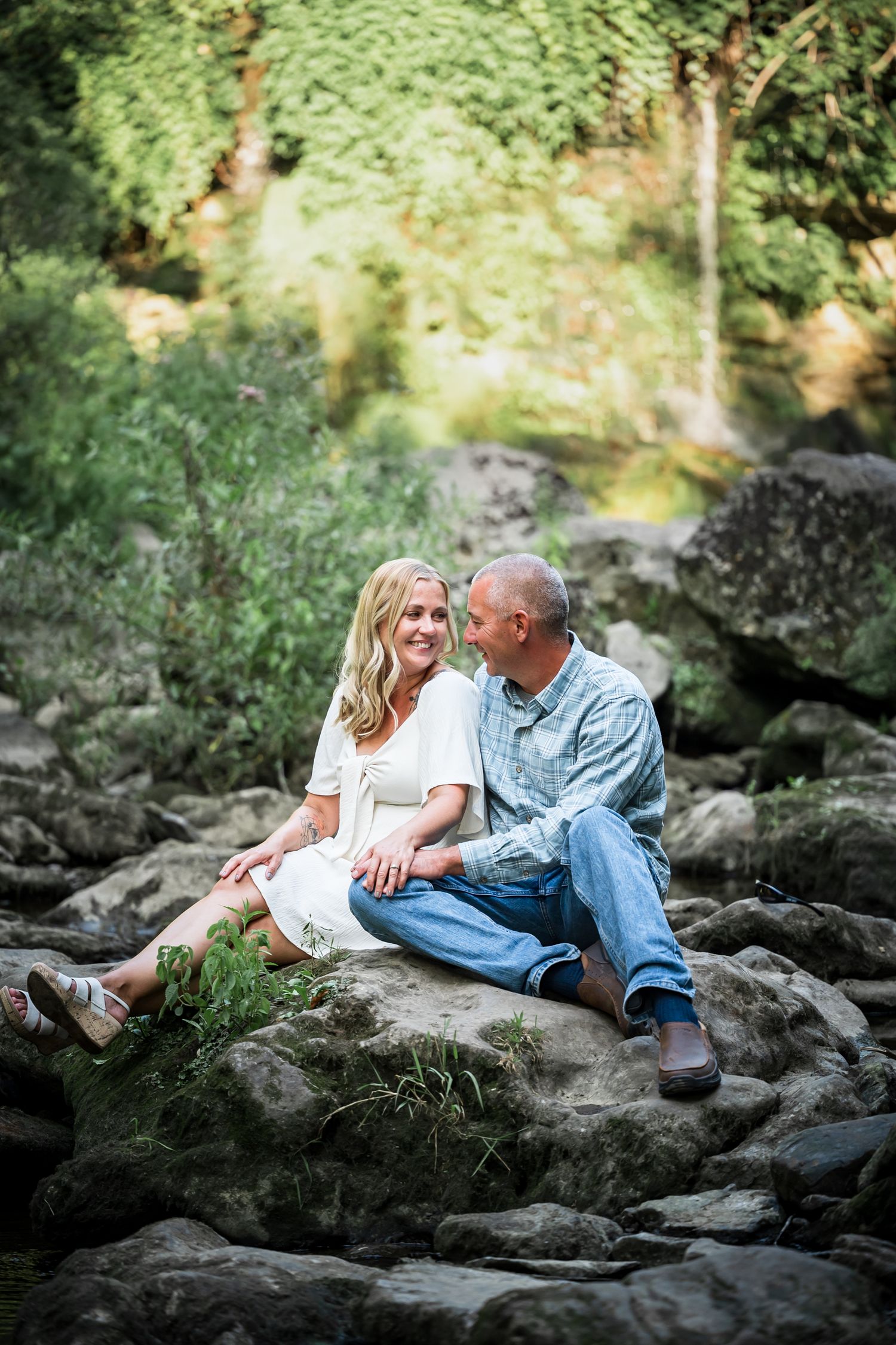 A couple sits on rocky outcroppings surrounded by green foliage near a waterfall.
