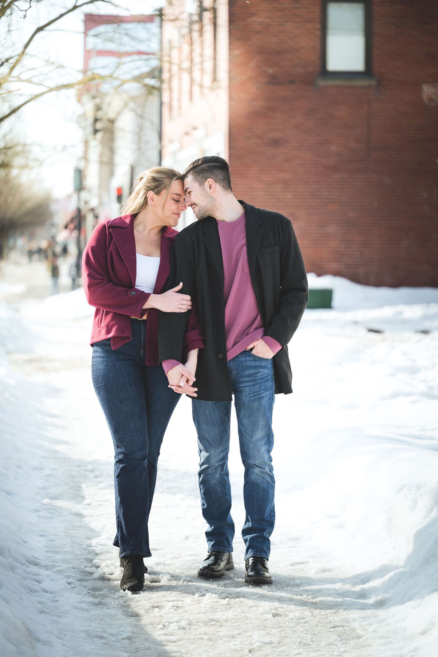 A couple shares an intimate moment while walking together on a snowy winter sidewalk near brick buildings.