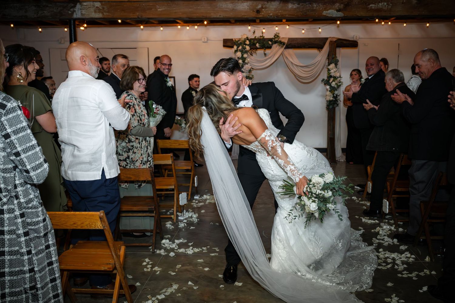 Wedding guests celebrate and dance together at a rustic indoor reception venue with string lights overhead.