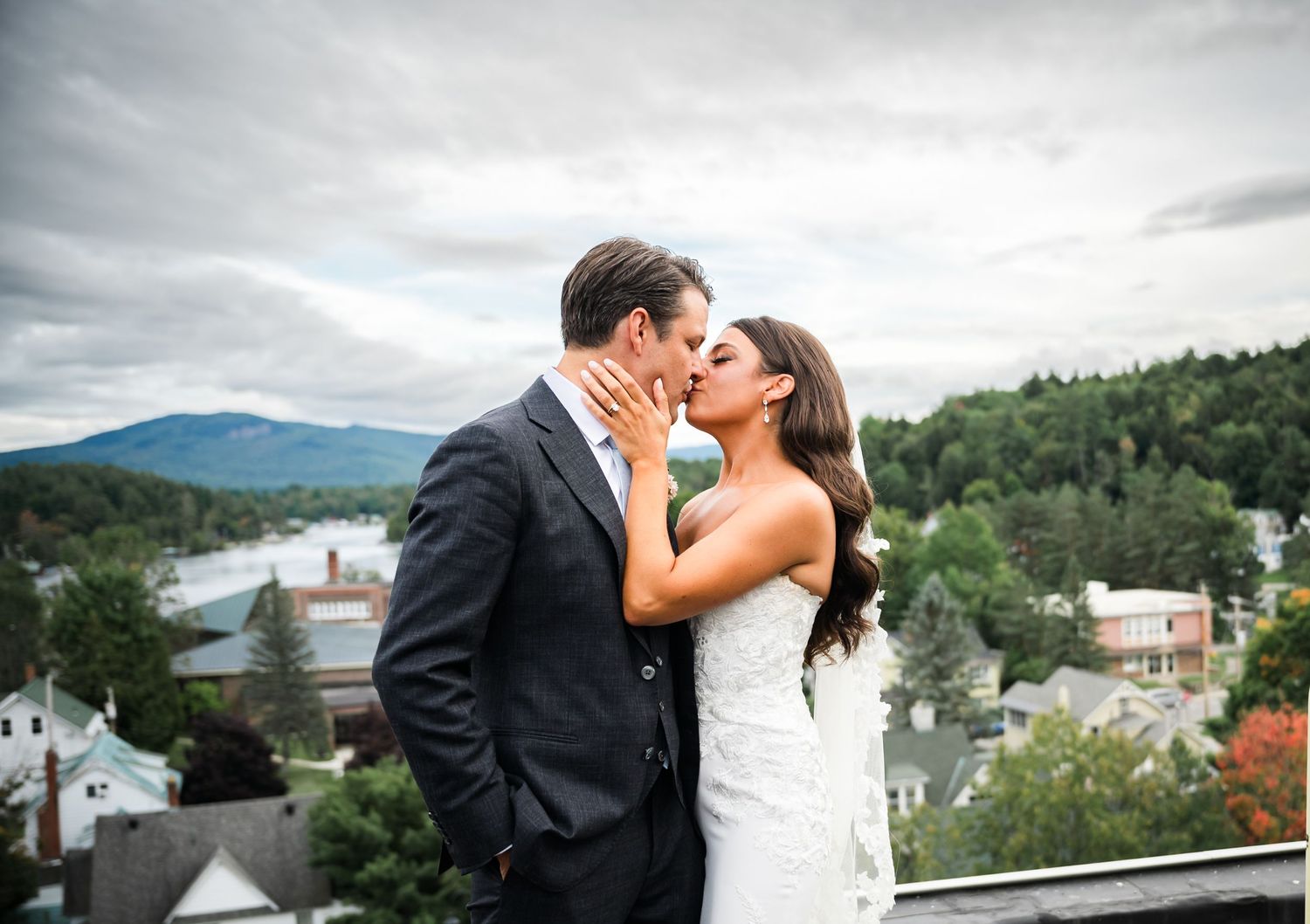 A romantic couple shares a tender kiss on a balcony overlooking a scenic mountain landscape and village below.