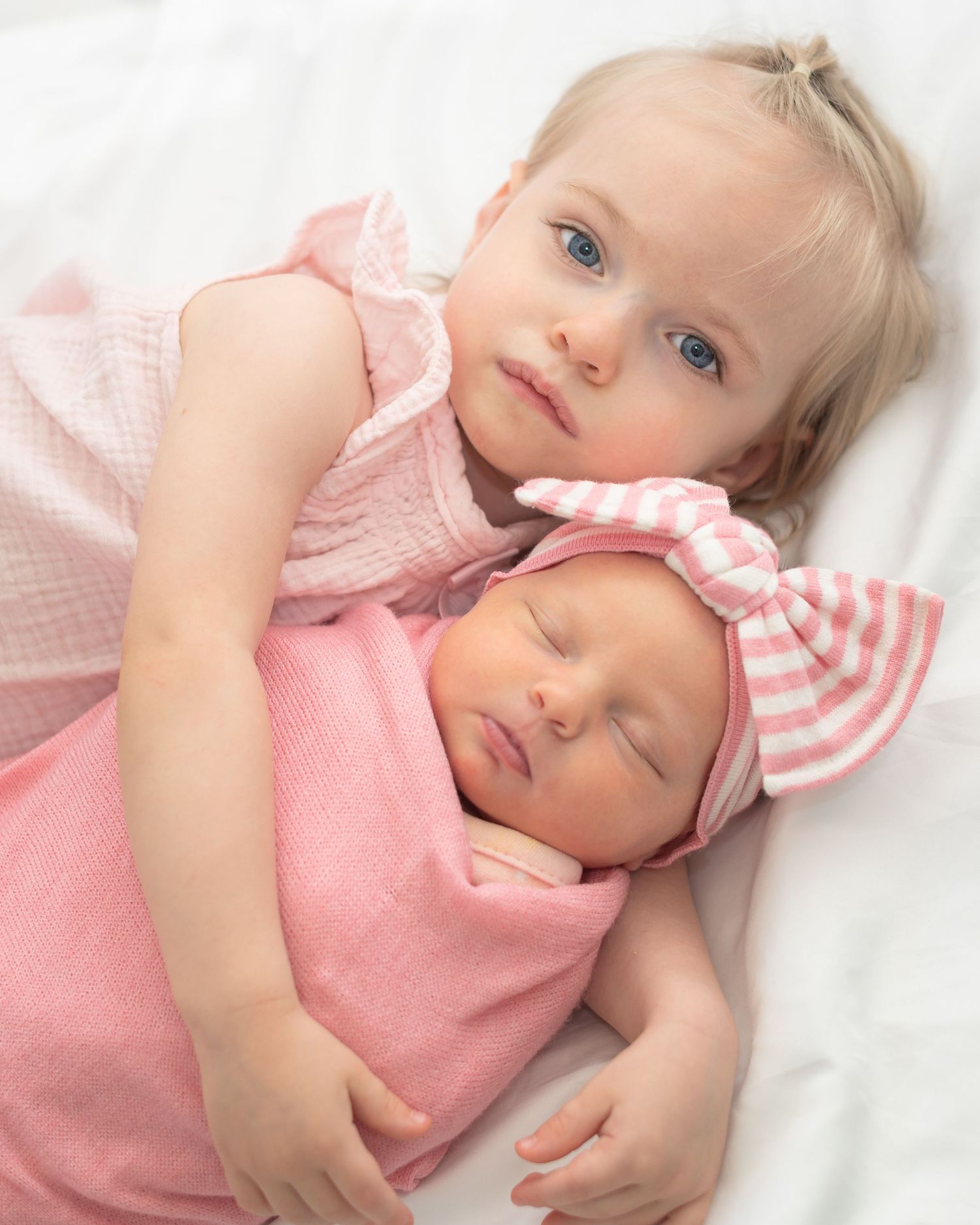 Two young children in pink outfits lying close together on white bedding while resting peacefully.