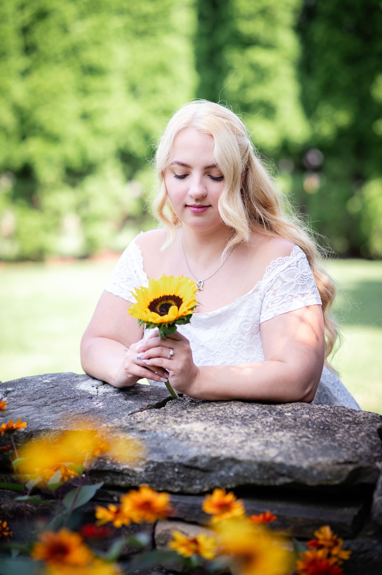 Woman in white lace dress holds a sunflower while sitting on stone wall in garden setting.