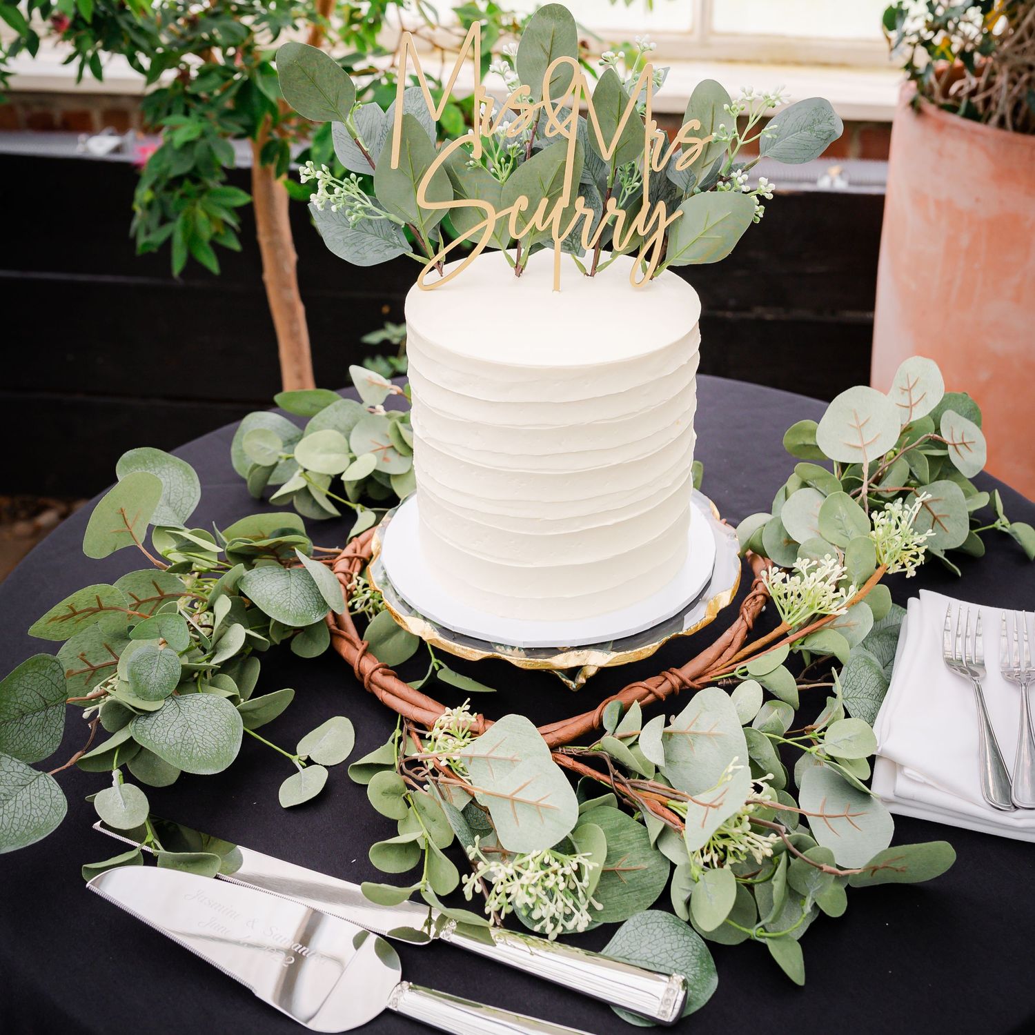 A white wedding cake decorated with eucalyptus leaves and a gold topper sits on a black table.