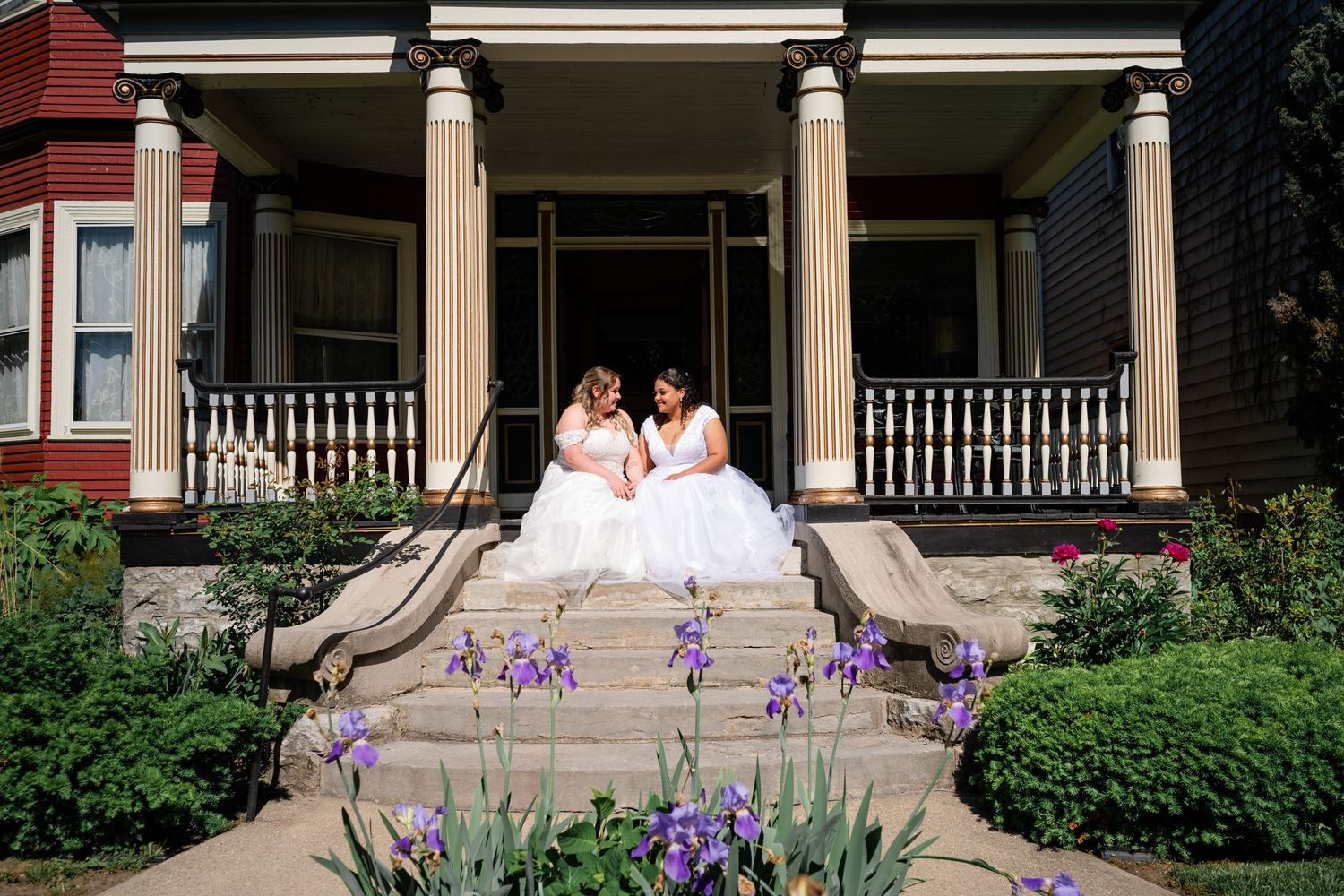 Two white gowns contrasting against a historic porch with purple flowers blooming in the garden.