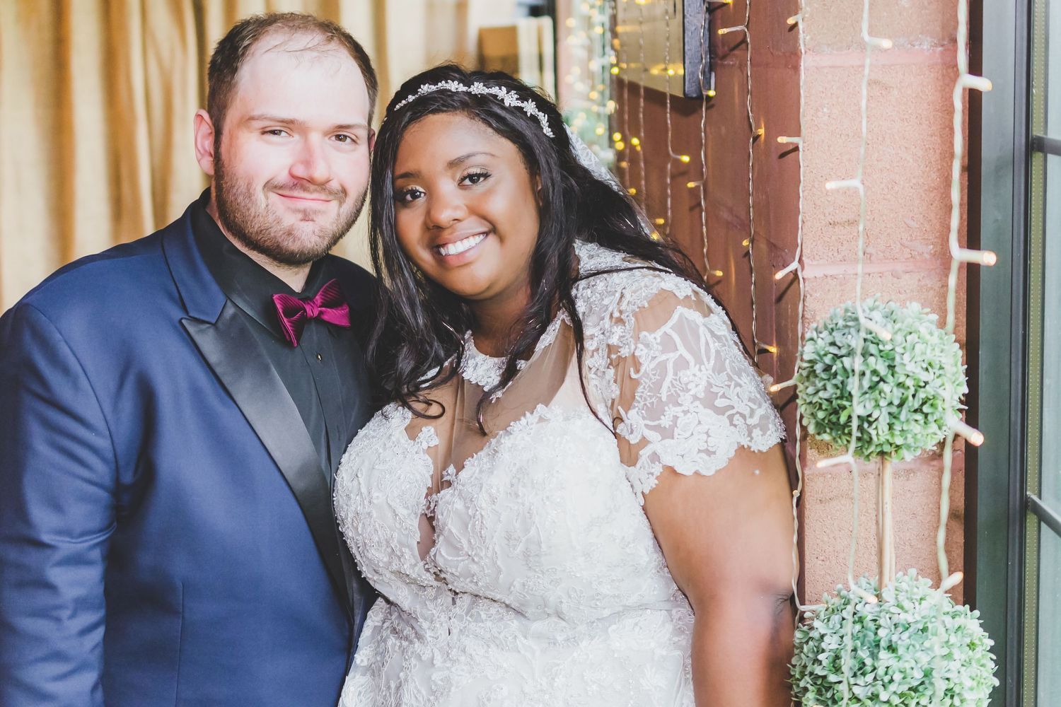 A wedding couple poses together against a backdrop with decorative greenery.