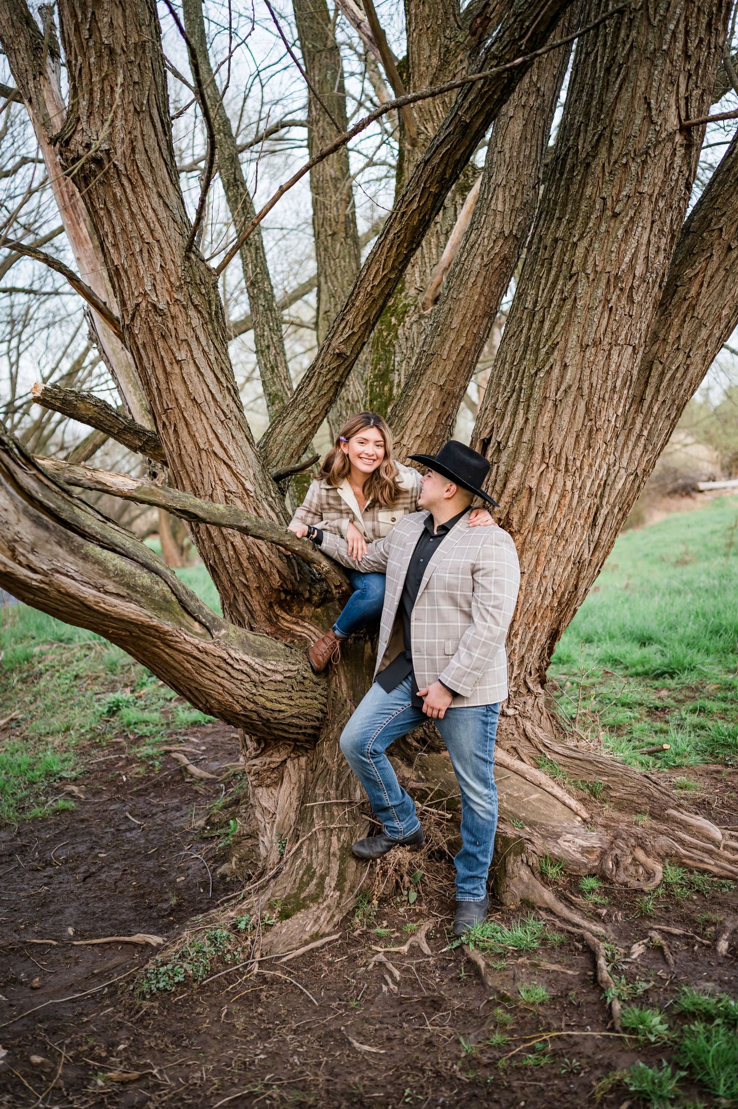 Couple poses playfully among branches of large old tree in countryside setting.