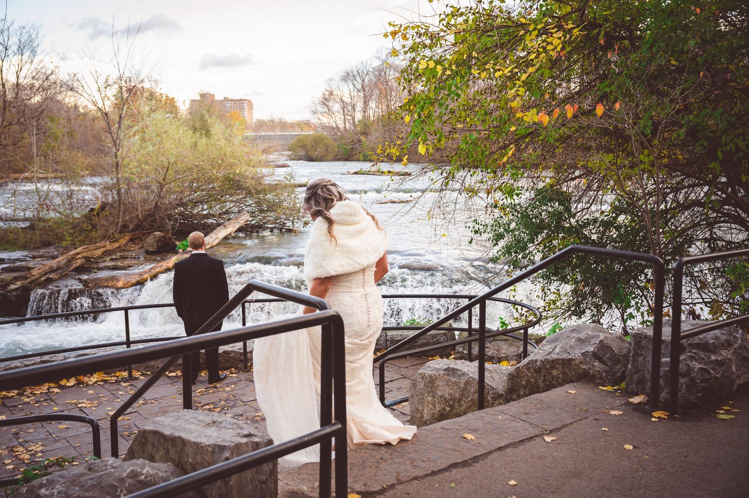 A couple stands on a stone observation deck overlooking rushing water and autumn scenery.