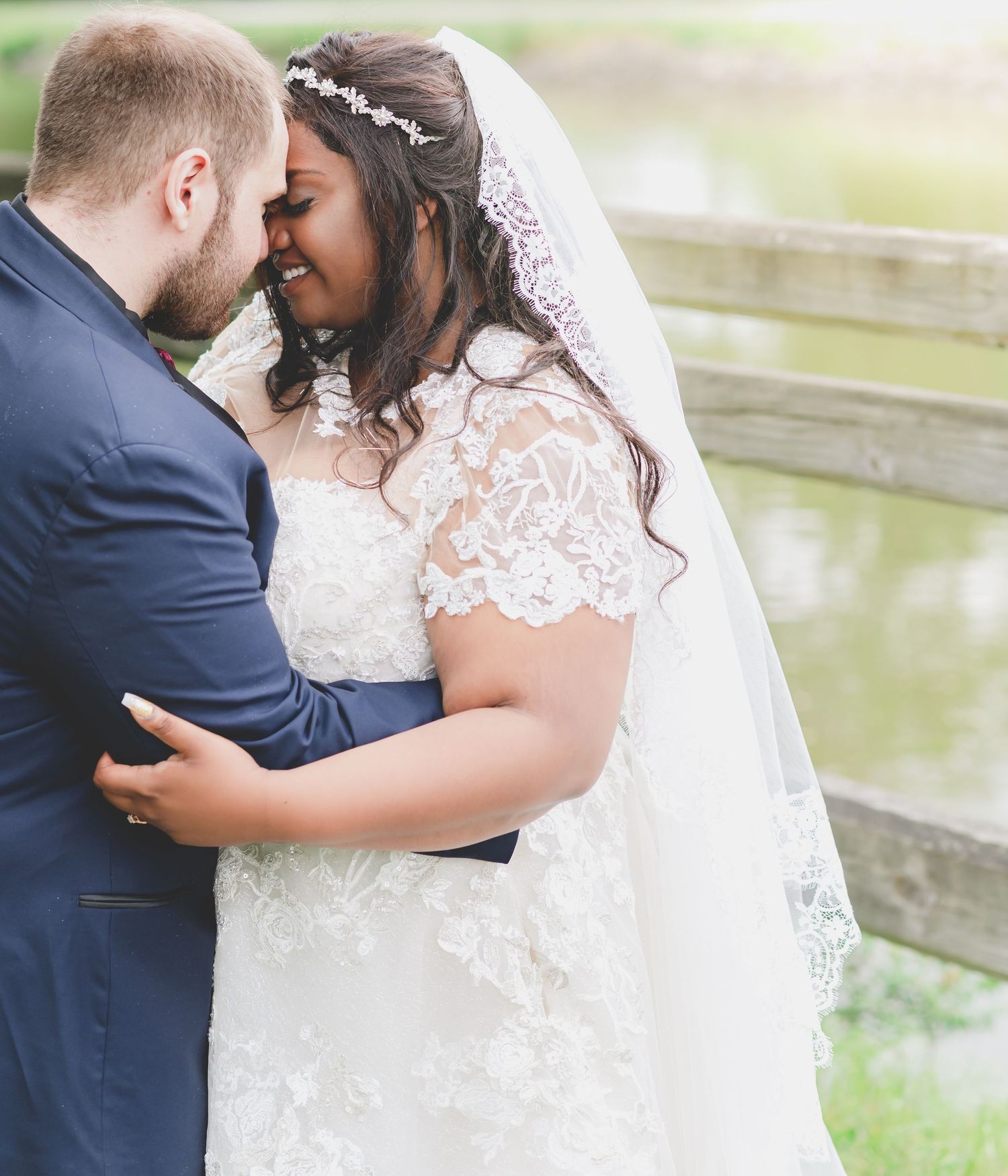 A tender moment between newlyweds embracing near a wooden fence by a peaceful pond.
