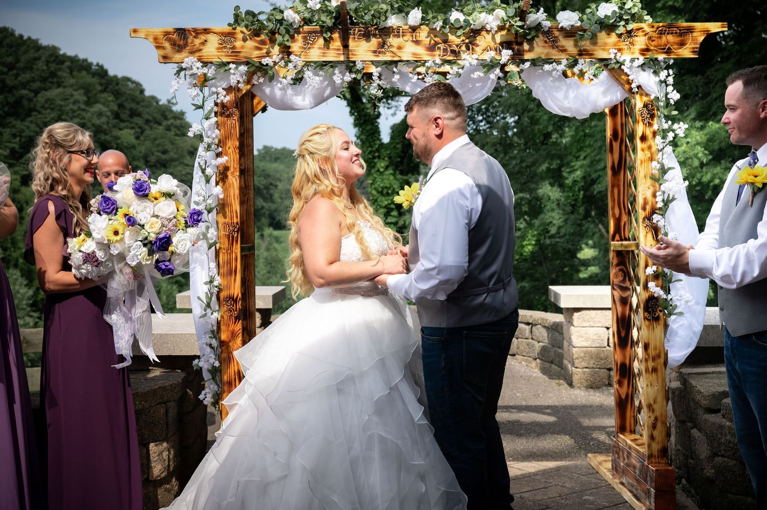 Wedding ceremony under wooden arch decorated with flowers and lights.
