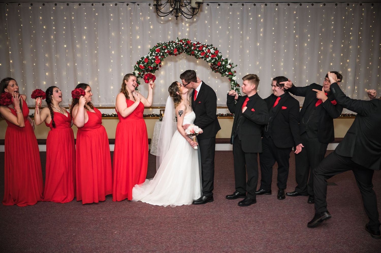 Wedding party reacting playfully during formal photos under a Christmas-decorated arch.