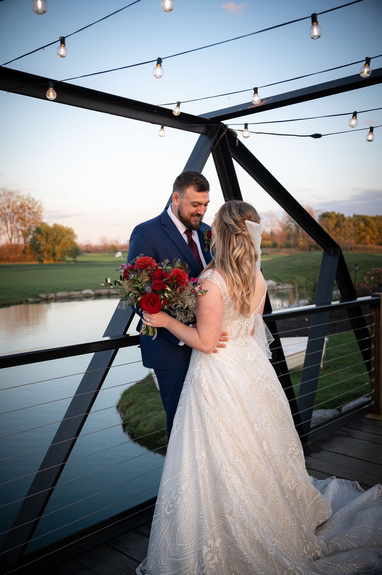 Romantic wedding couple on deck with string lights and red bouquet at sunset over water.