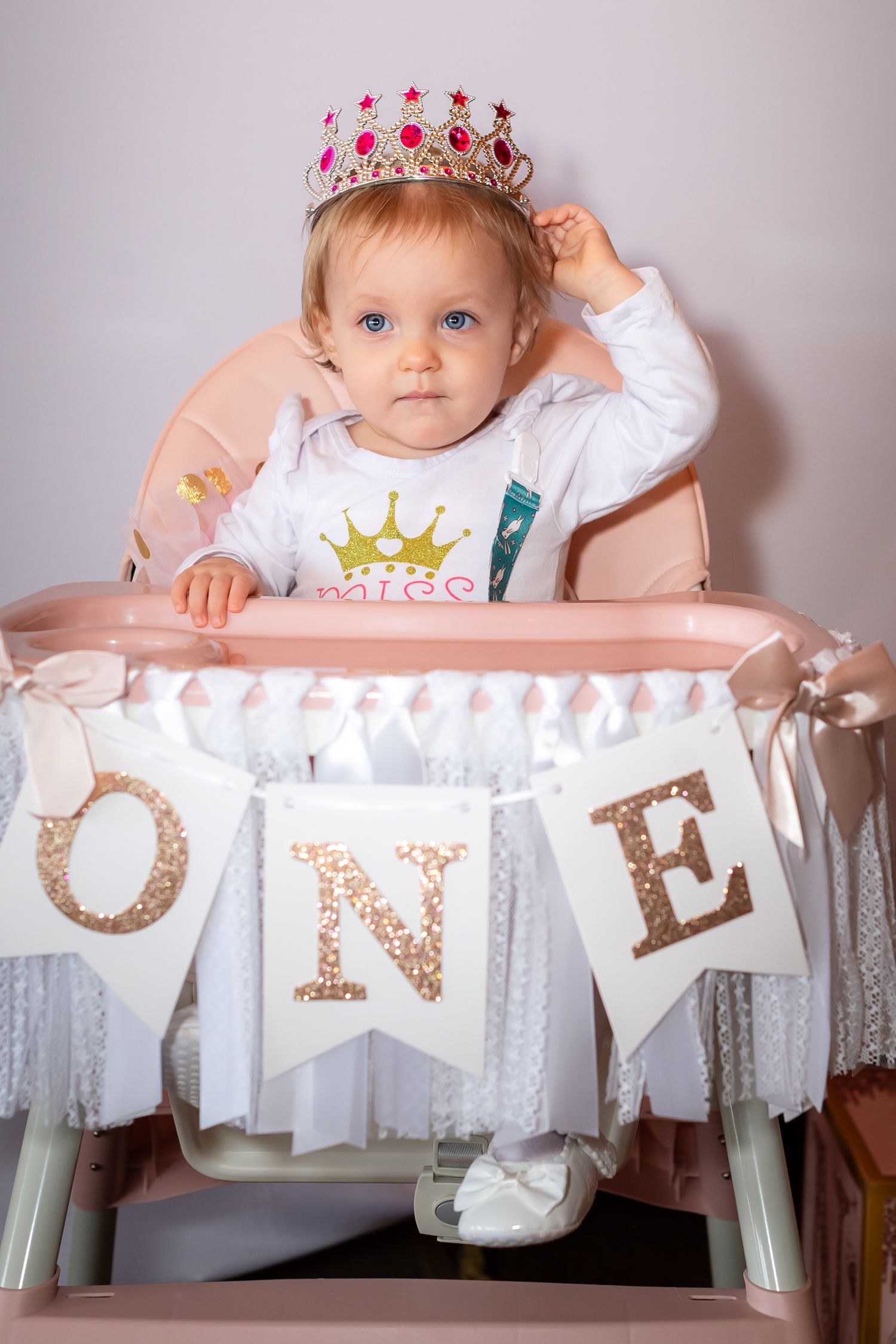 Baby in high chair with decorative 'ONE' banner celebrating first birthday milestone with crown and pink details.