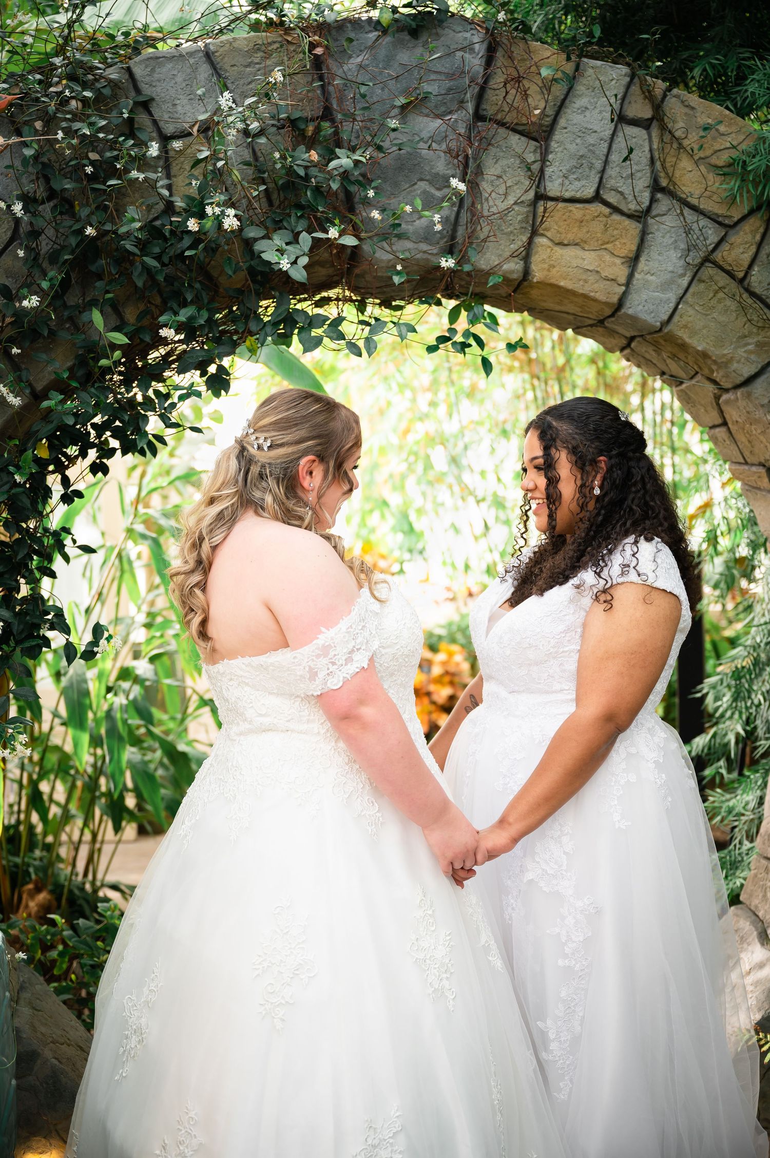 Two brides in white wedding dresses hold hands while standing under a stone archway with lush greenery.