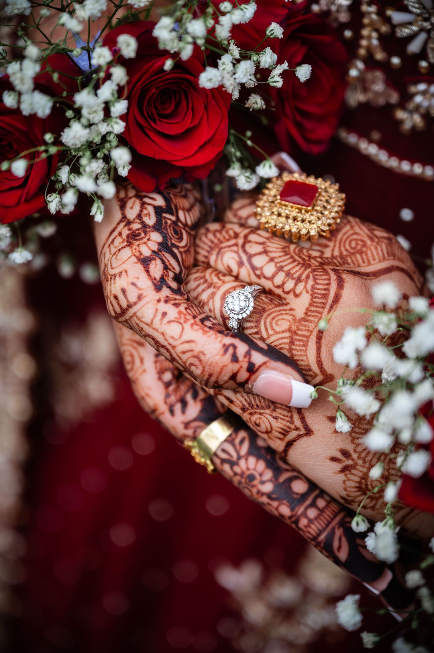 Intricate henna design adorns a hand with red roses and baby's breath flowers against a dark red background.