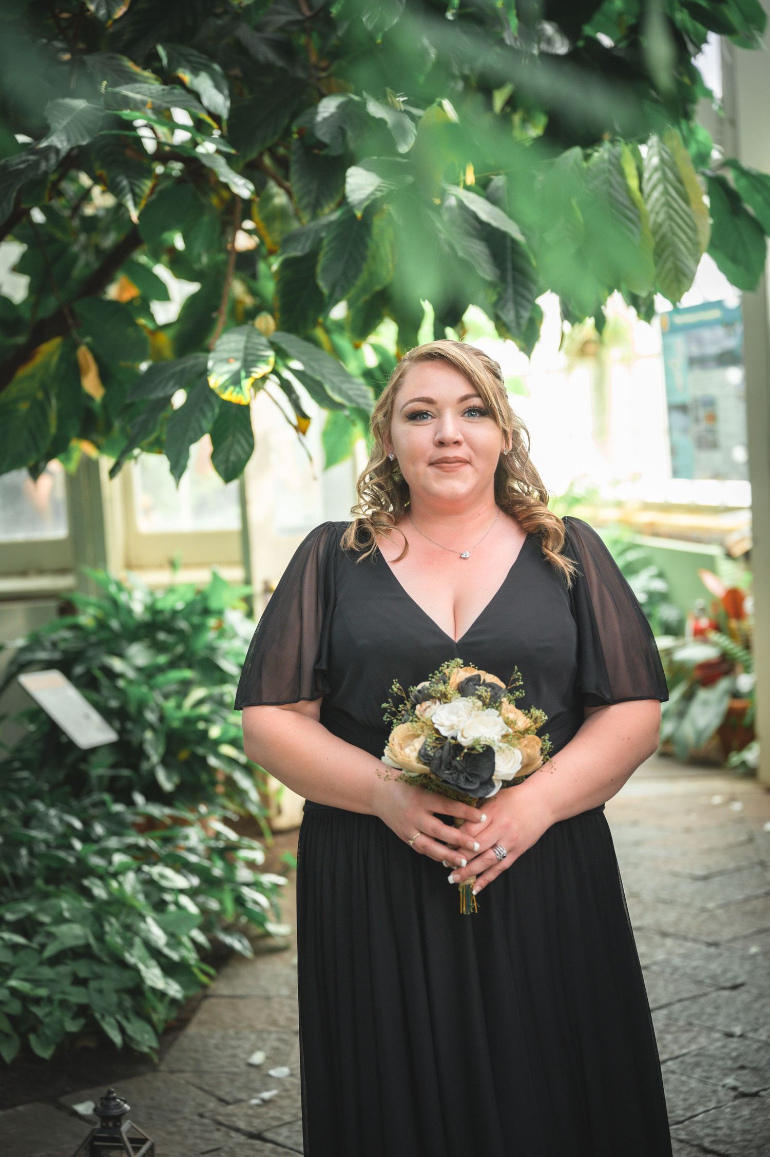 Bridesmaid in black dress with sheer sleeves holds flower bouquet under garden foliage.