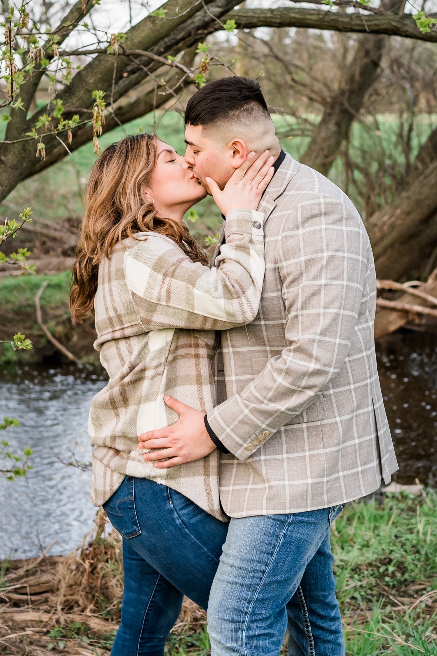 Couple sharing intimate moment by flowering tree near stream wearing coordinated beige plaid outfits.