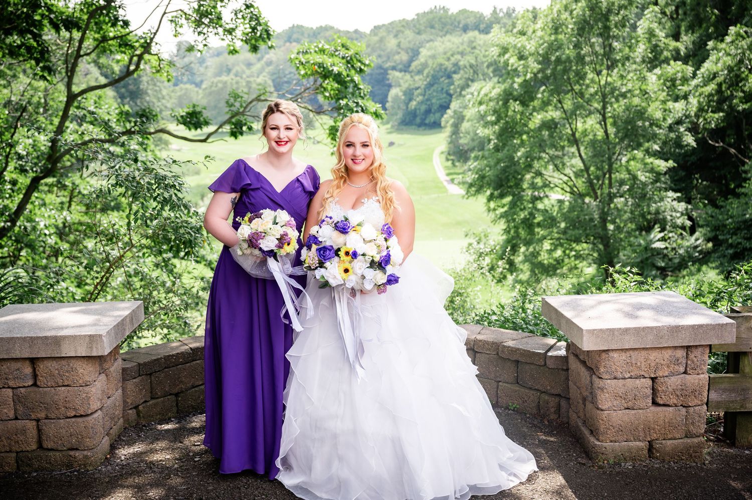 Two people in formal attire holding purple and white bouquets pose on a stone terrace with scenic view.