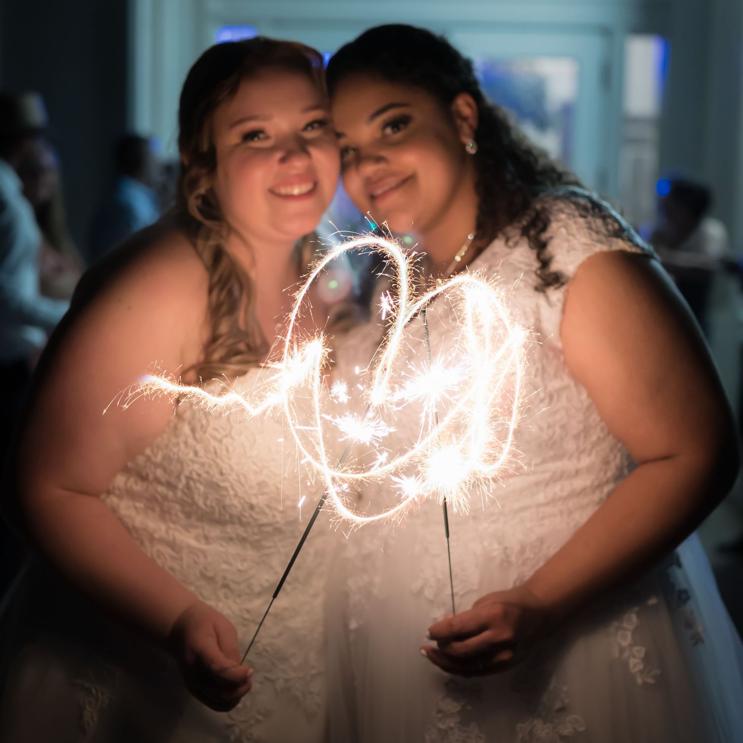 Two brides create heart shape with sparklers in dark setting.