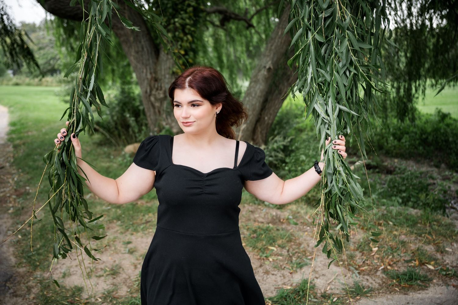 Standing between willow tree branches in a black dress outdoors.