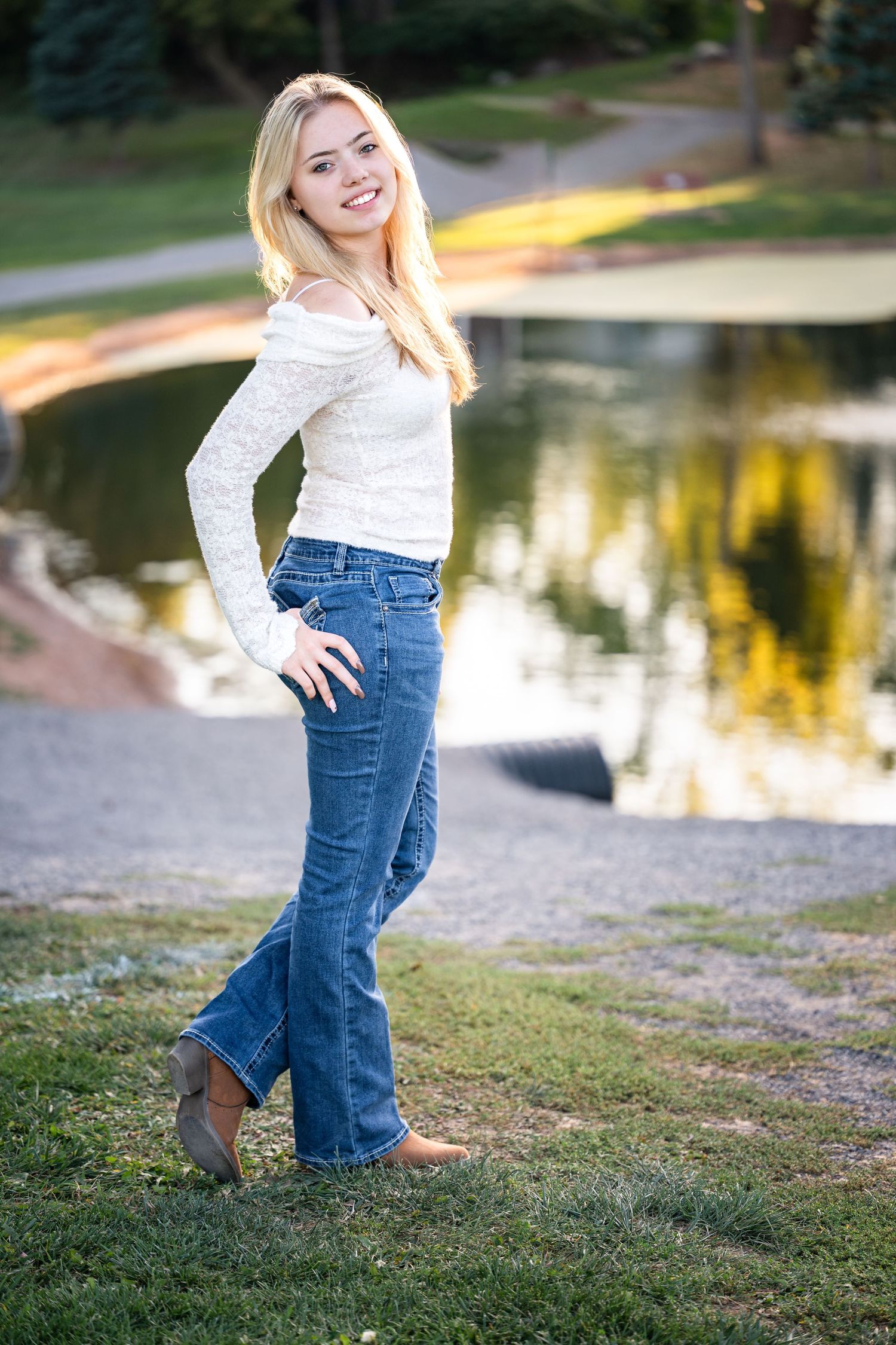 Child in white sweater and jeans plays by a pond at sunset with autumn reflections in the water.