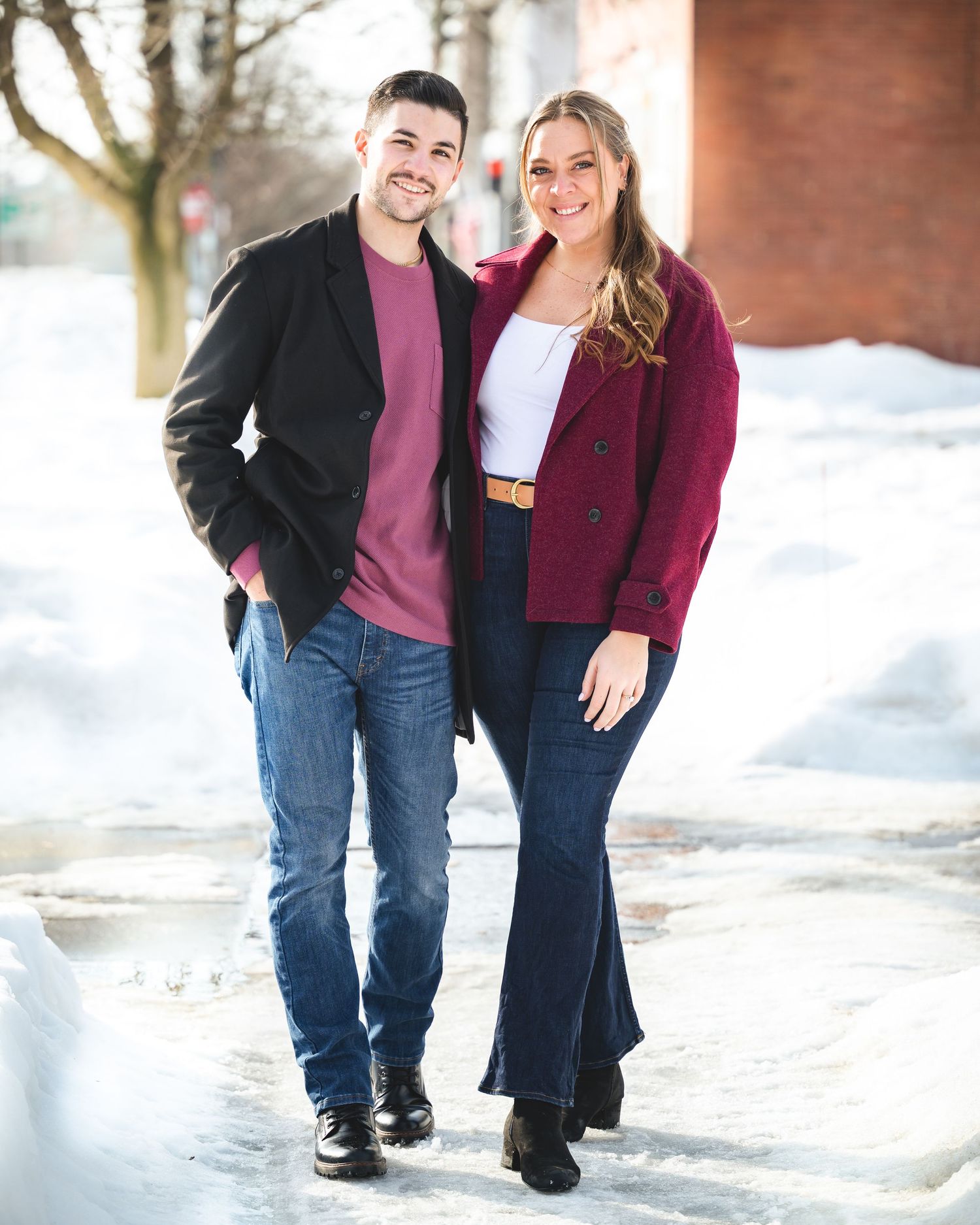 A smiling couple poses together outdoors in winter, wearing coordinating burgundy and black outfits against a snowy backdrop.