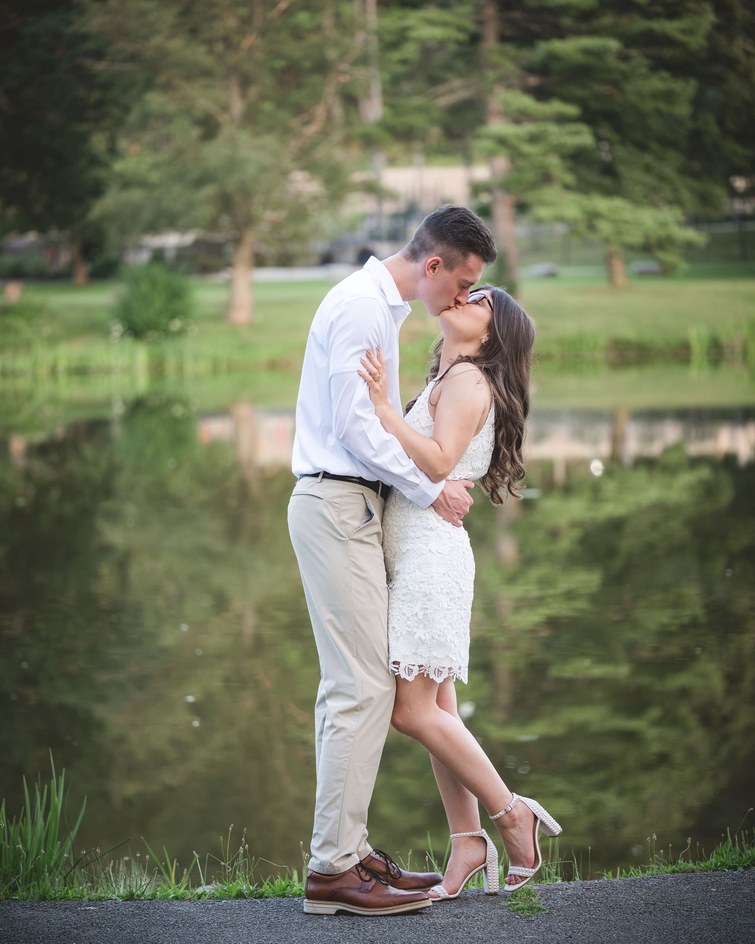 Couple sharing a romantic kiss by a serene pond, man in white shirt and khakis, woman in white lace dress and heels.