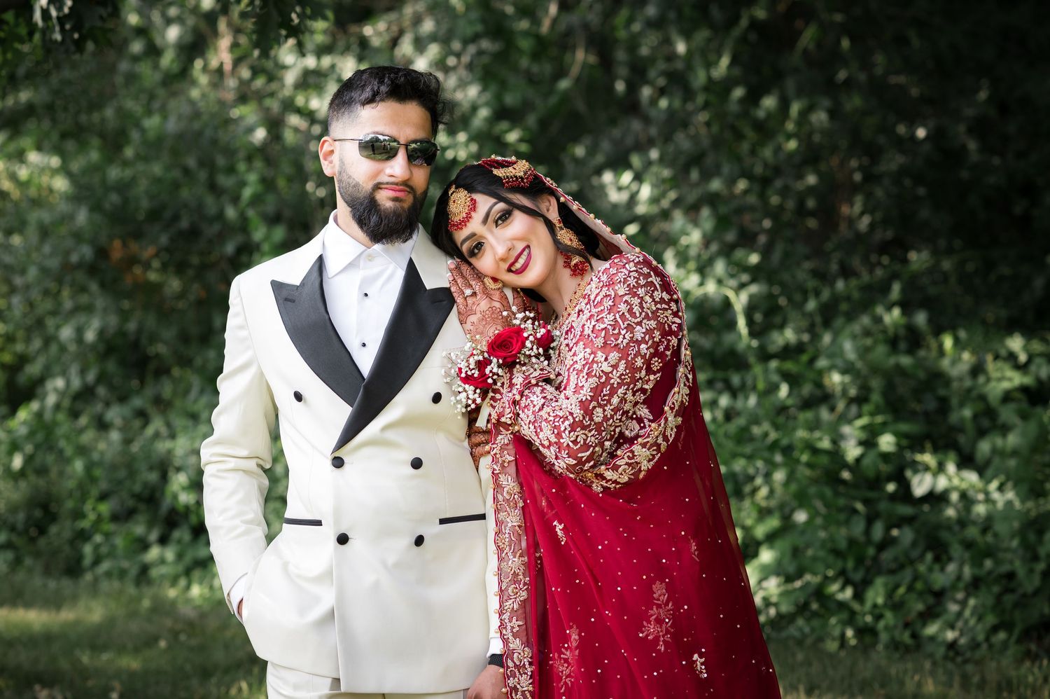 A couple in traditional South Asian wedding attire featuring a red bridal outfit and white double-breasted suit standing outdoors.