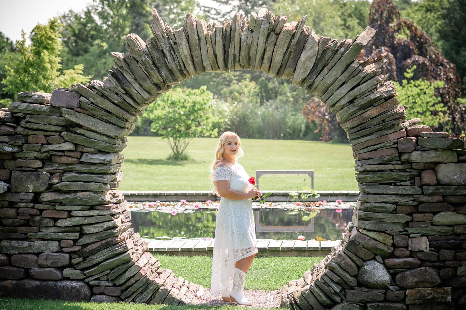 A bride poses in a white dress within a circular stone archway in a garden setting.