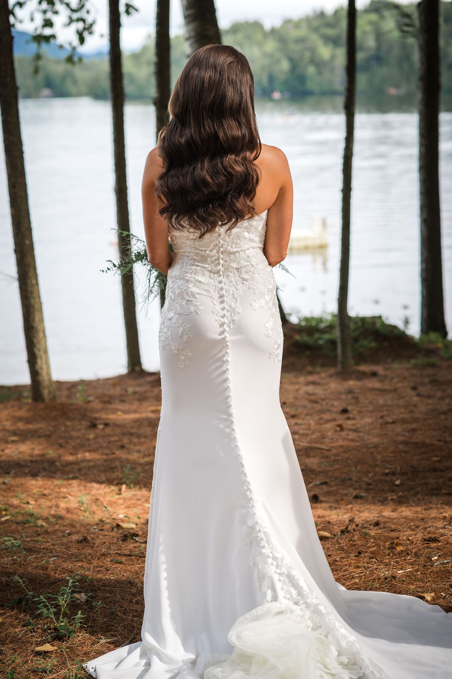 Back view of a bride in a fitted lace wedding dress with button details standing by a lake.