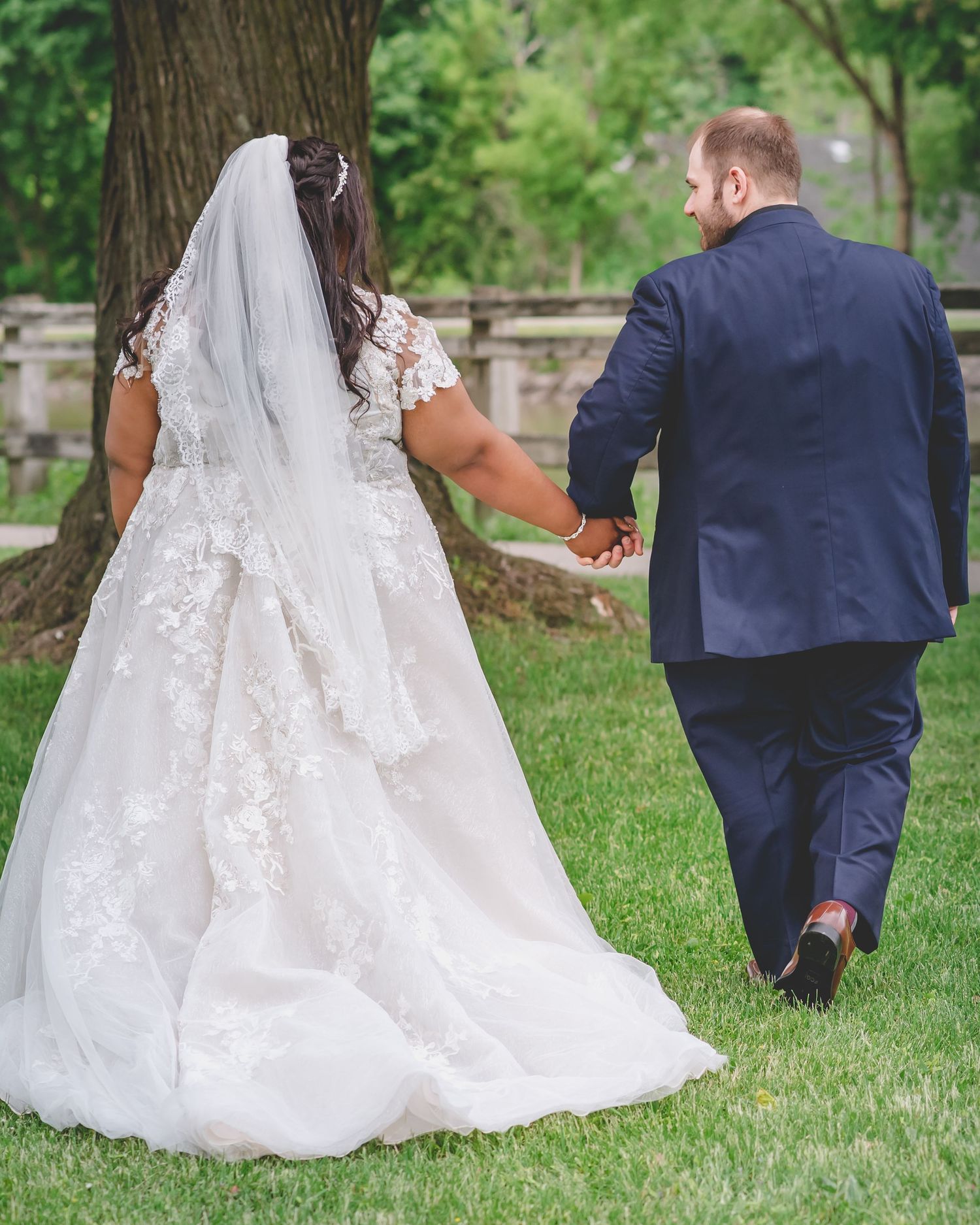 Wedding couple walks hand in hand on grassy path with flowing white dress trailing behind.