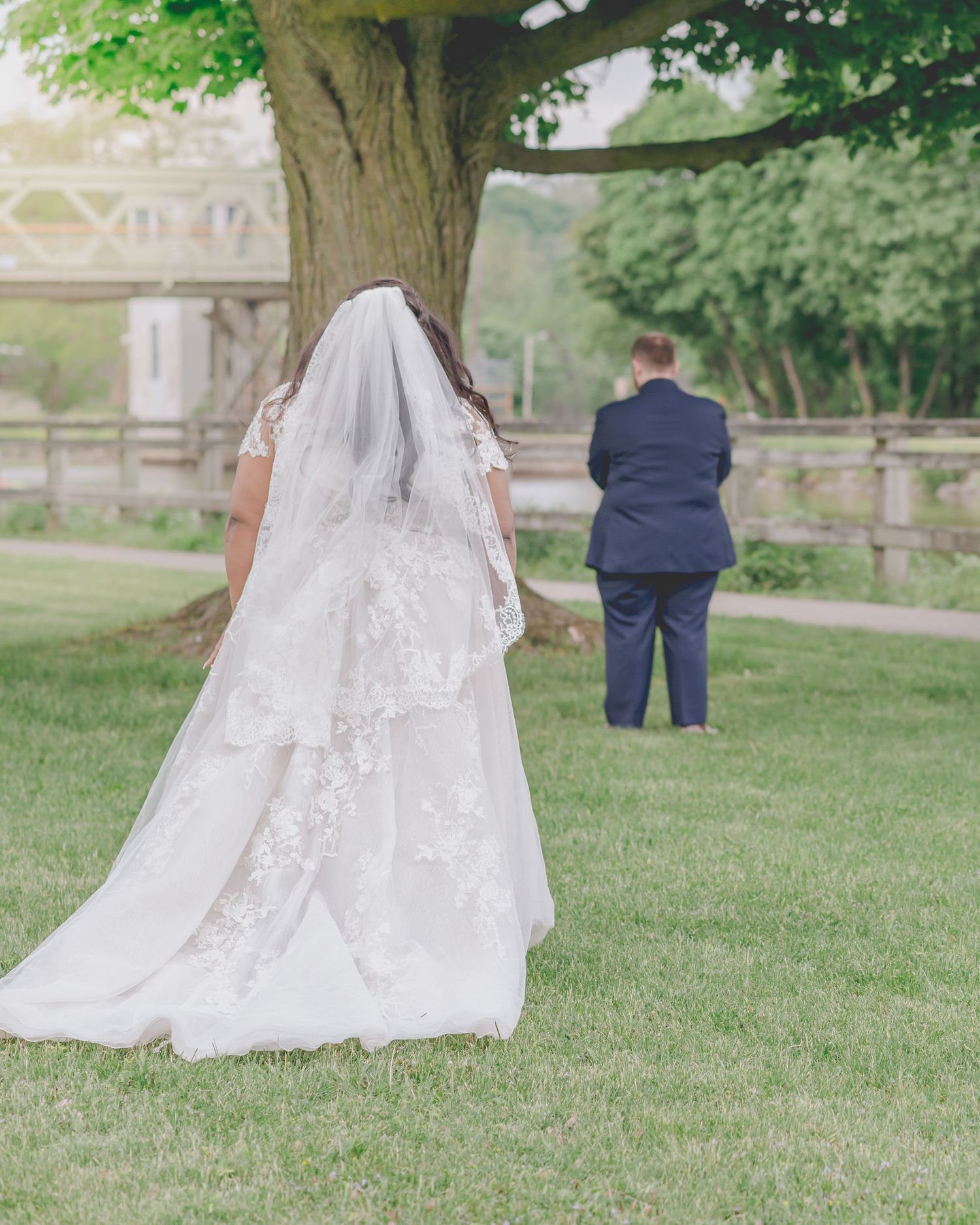 A bride in a flowing white gown stands near a tree during a first look moment.